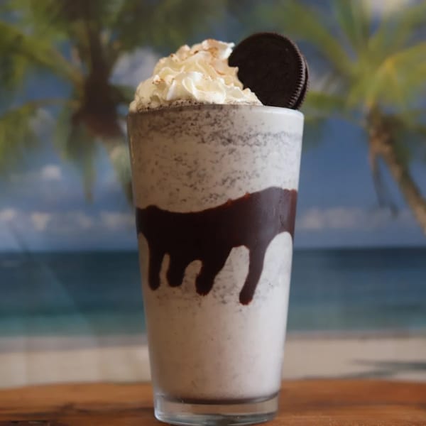 Oreo milkshake with whipped cream and chocolate drizzle, set against a beach and palm tree background.