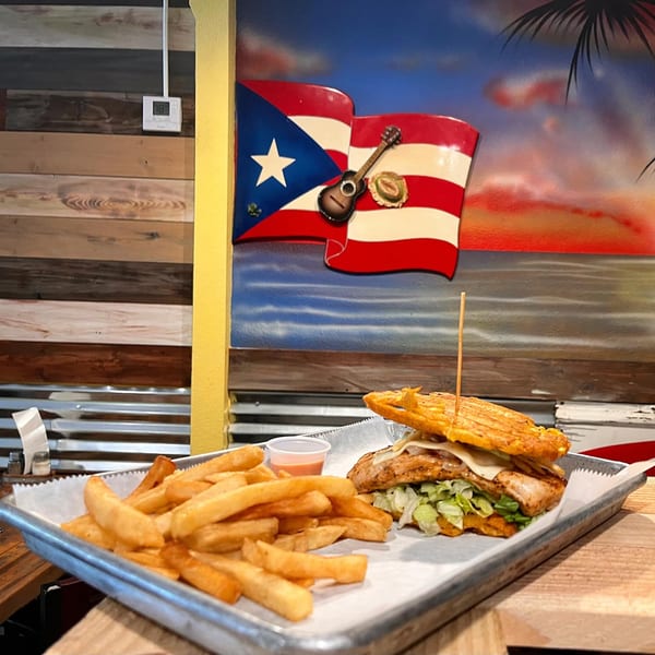 A Puerto Rican sandwich with fries on a tray, against a mural of the Puerto Rican flag.