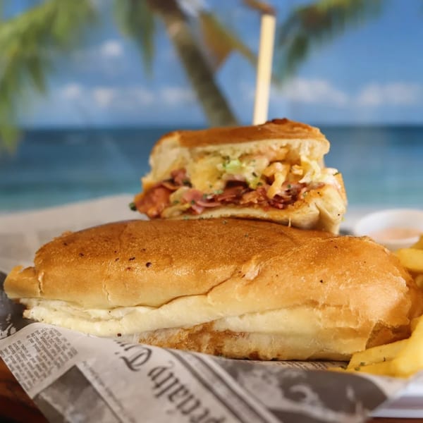 A Puerto Rican sandwich with meat and cheese, served with fries on newspaper, against a beach backdrop.