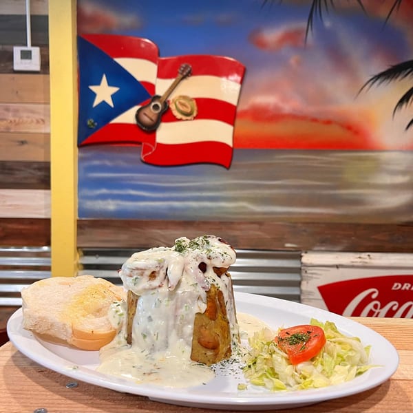 Mofongo with creamy sauce, garlic bread, and salad, against a Puerto Rican flag mural.