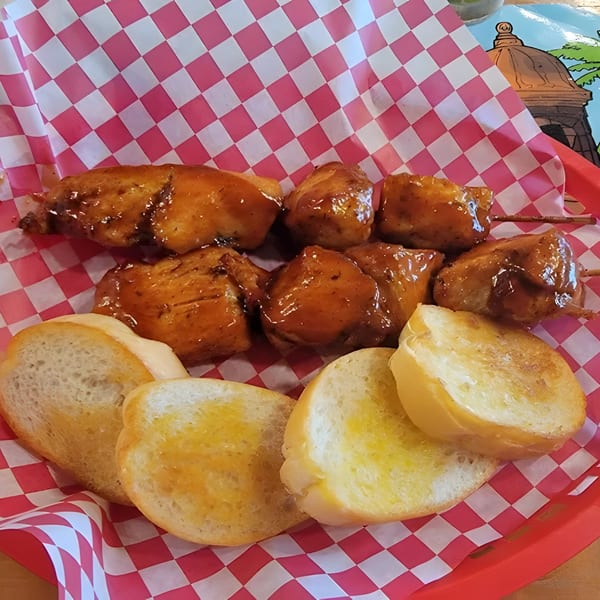 Two skewers of glazed chicken pinchos with toasted bread slices in a red checkered basket.