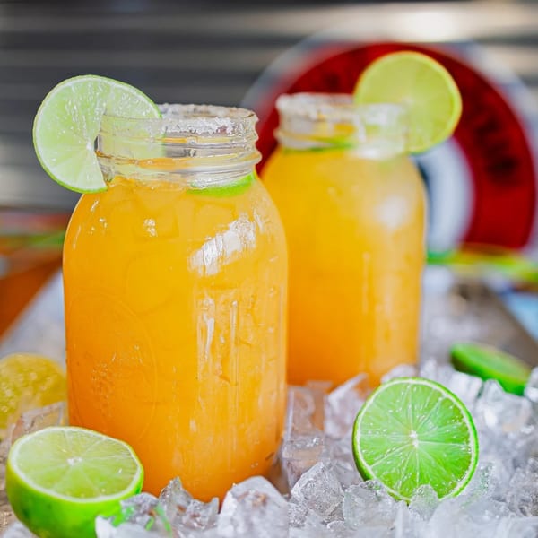 Two mason jars of orange drinks with lime slices and salted rims, surrounded by ice.